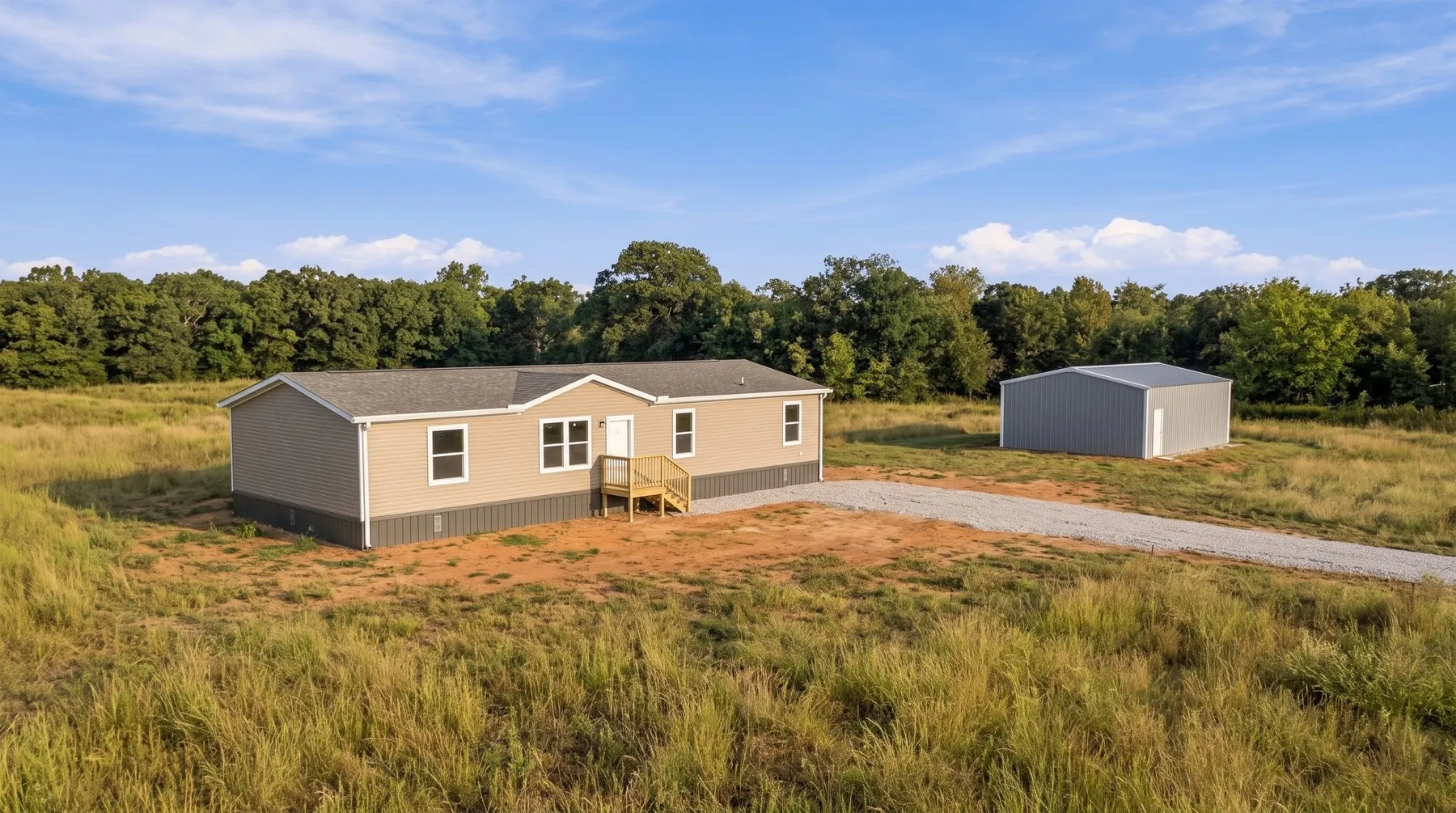 A new single-storey house sitting small in a wide open pasture, with a metal workshop building beside it and an oak treeline behind.